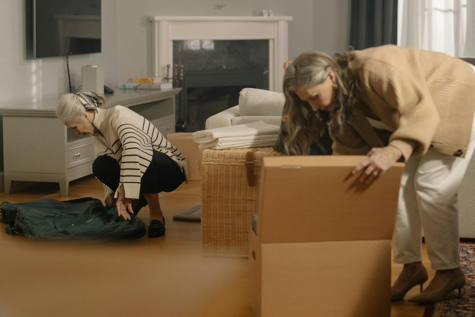 Two senior women packing belongings in boxes, preparing for a move in a cozy living room.